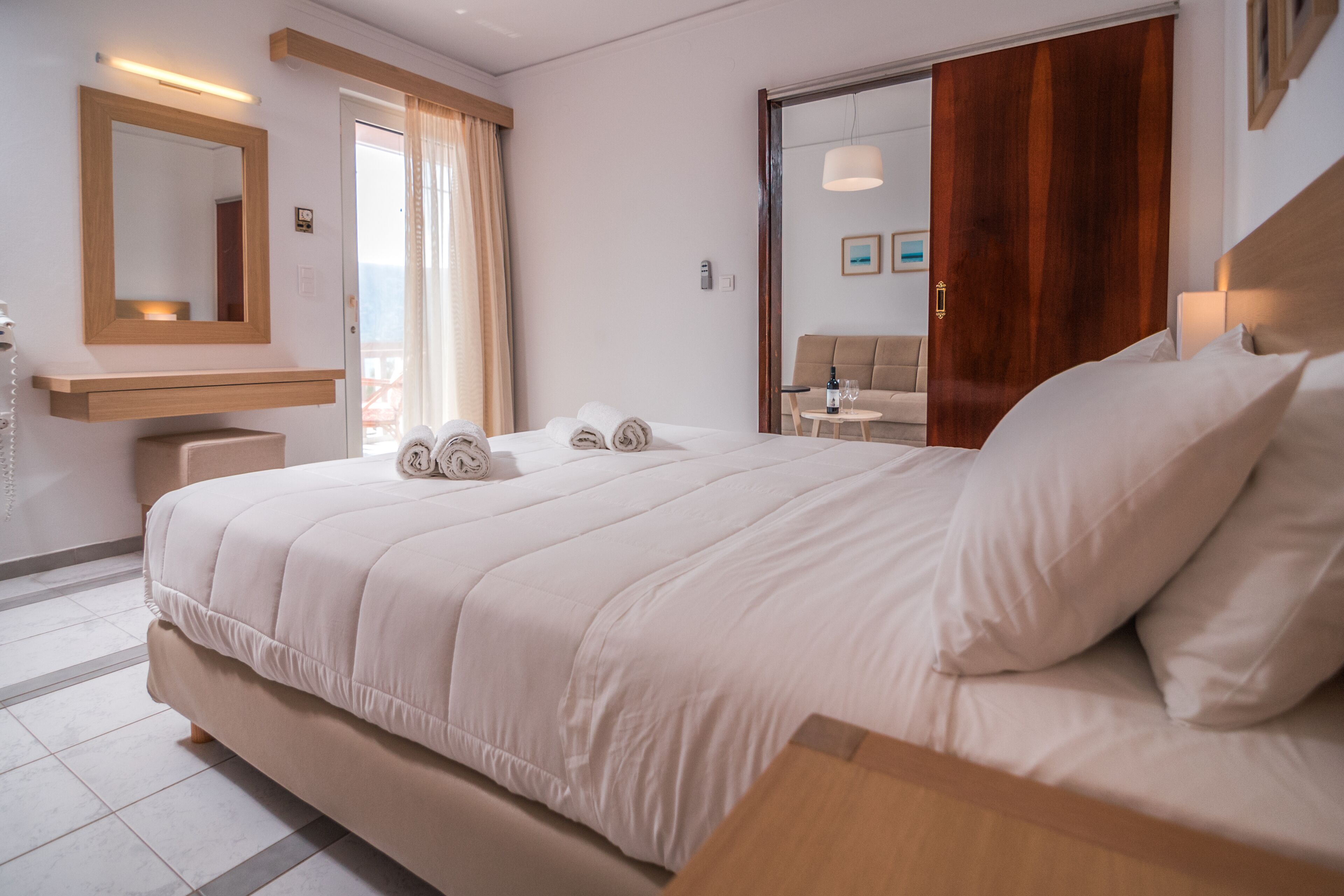 Una habitación de hotel con una cama grande, cabecera de madera, un espejo, una mesita de noche y vistas al balcón.