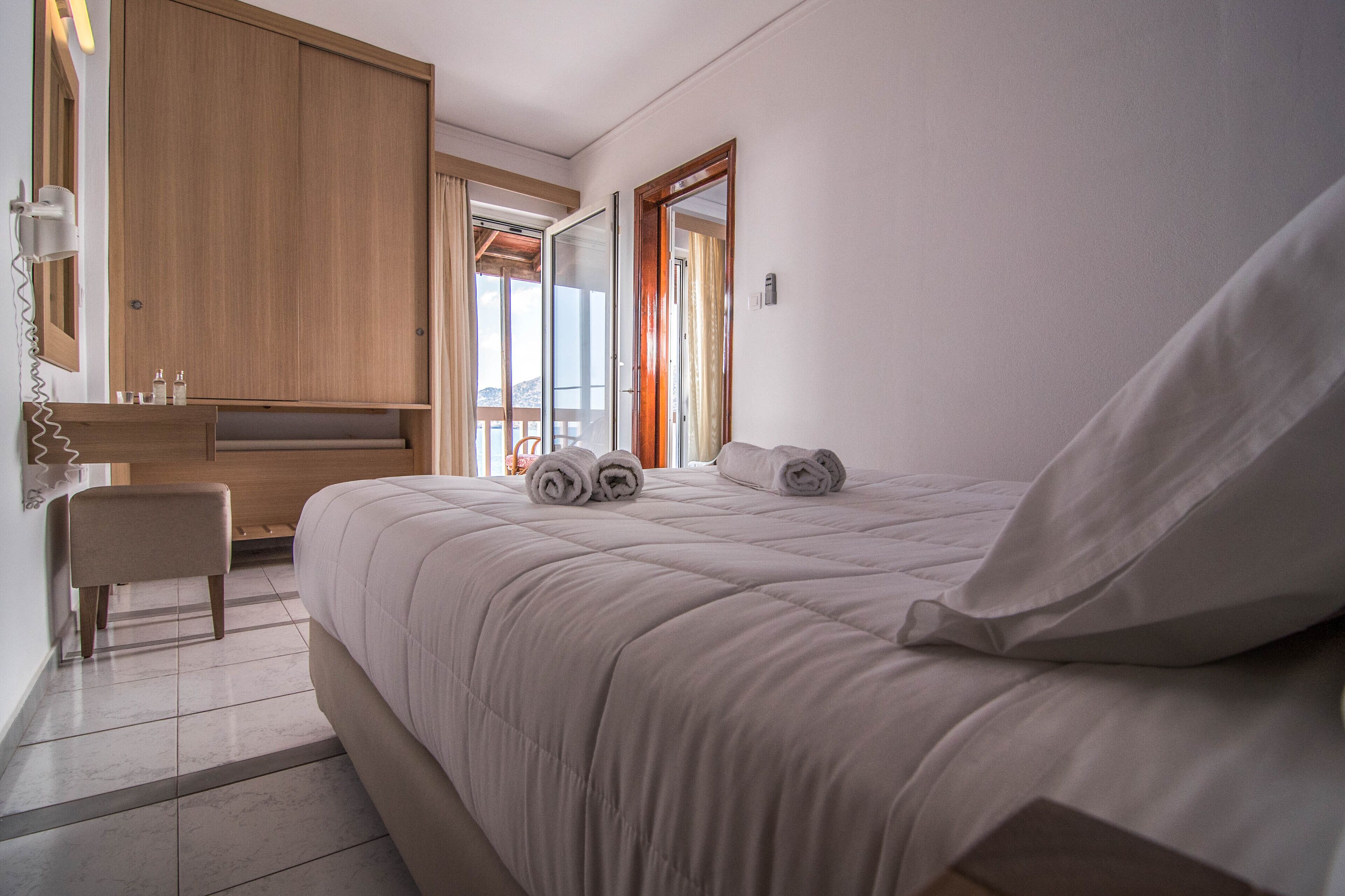 A bedroom with a bed, a wooden wardrobe, a bench, and a bathroom with a mirror and towel rack.