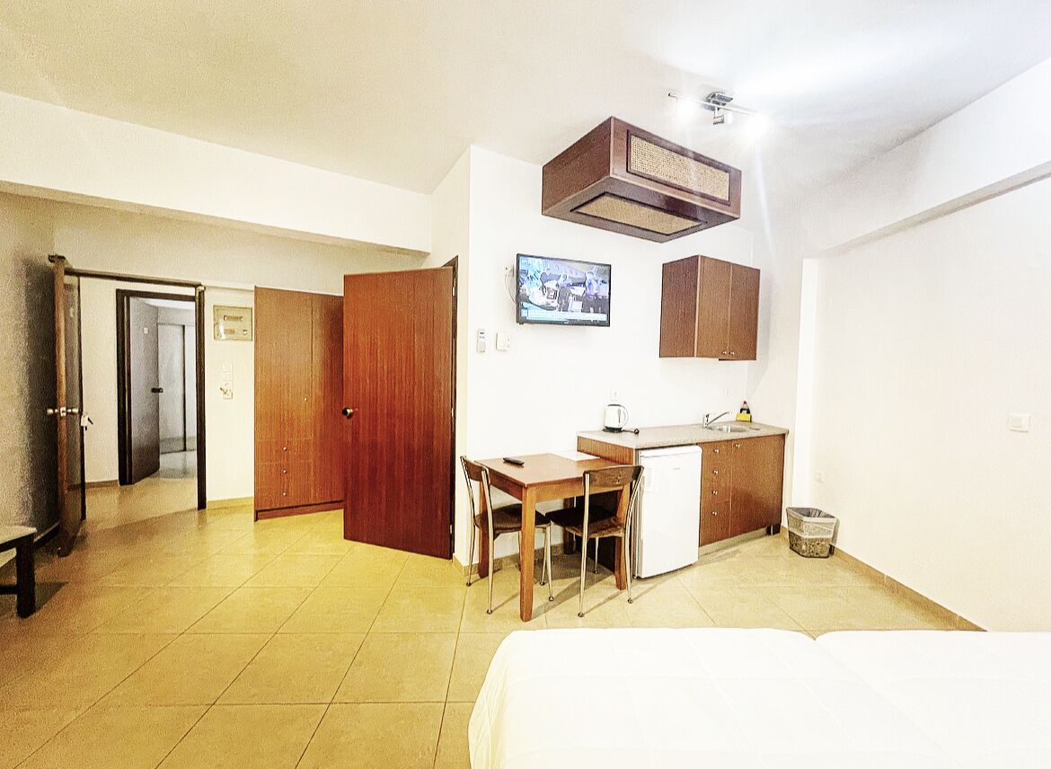 A compact hotel room with a bed, a small kitchenette, a wooden desk, and a television.