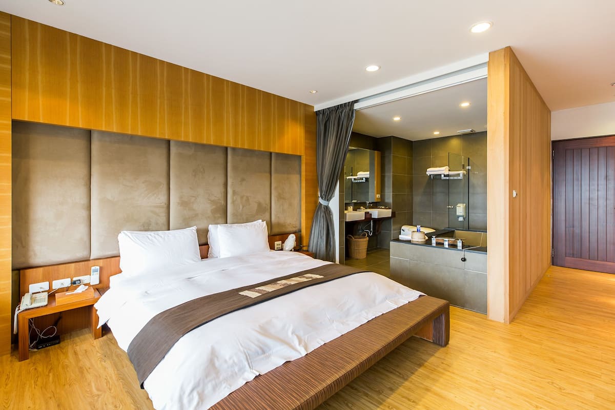 A modern hotel room with a large bed, wooden headboard, and a bathroom visible through an open door.