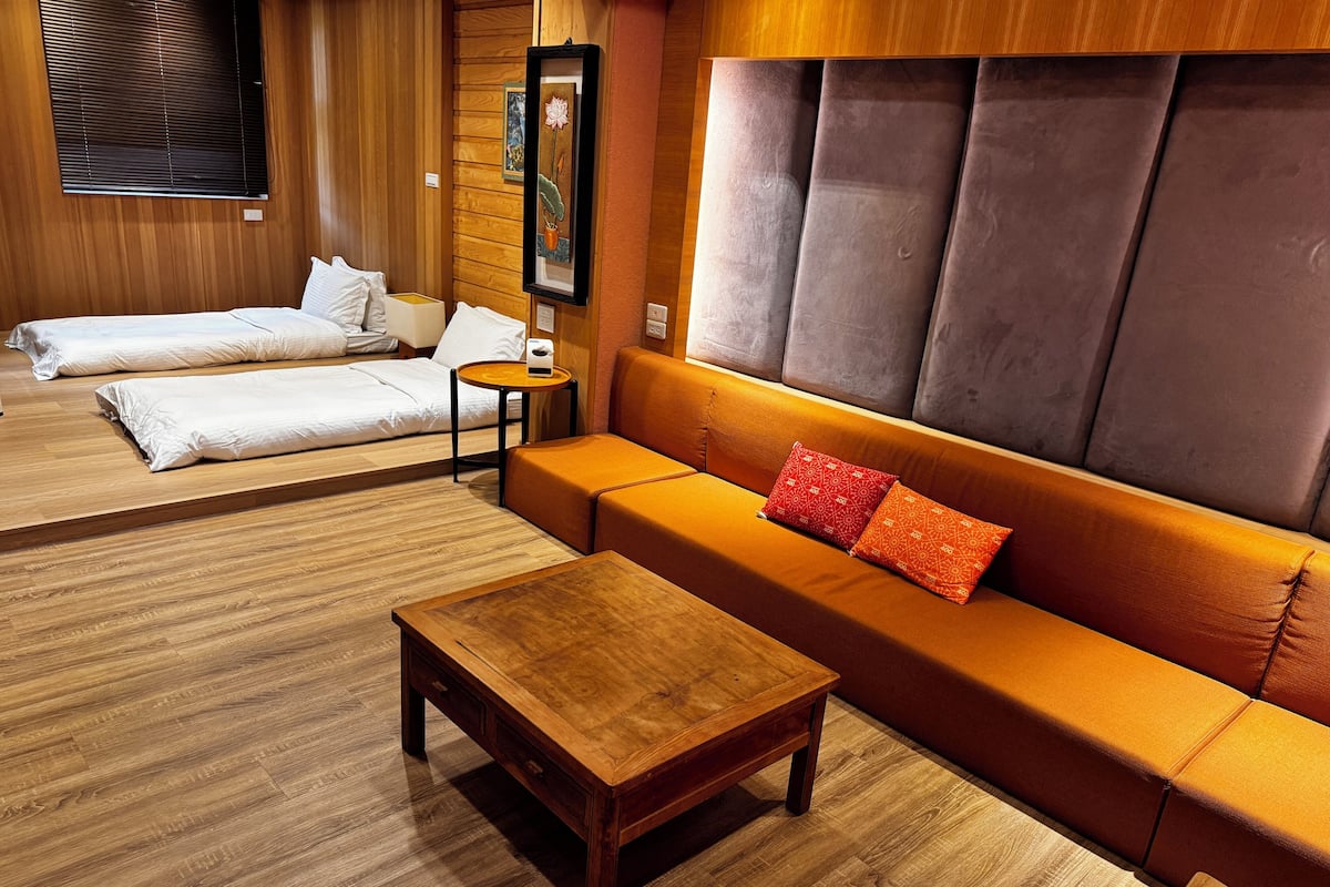 A room with a wooden floor, a sofa with orange cushions, a wooden coffee table, and a bed with white linens.