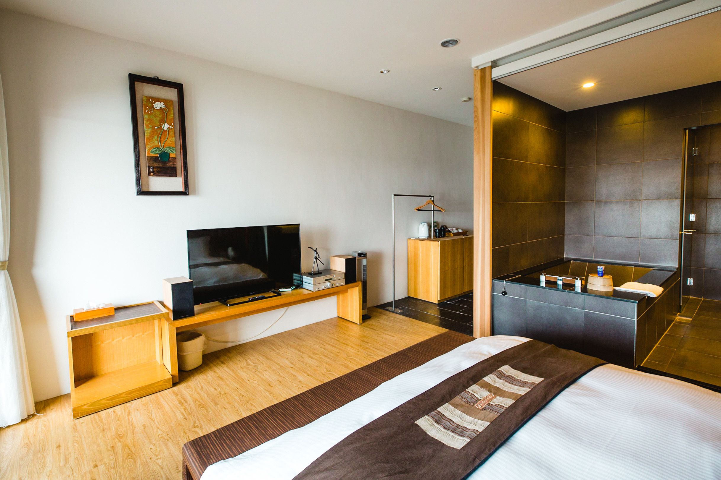 Ein modernes Hotelzimmer mit einem großen Bett, einem Fernseher, einem Holznachttisch, einem kleinen Schreibtisch, einem Spiegel, einem Kleiderschrank und einem Badezimmer mit Dusche und Waschbecken.