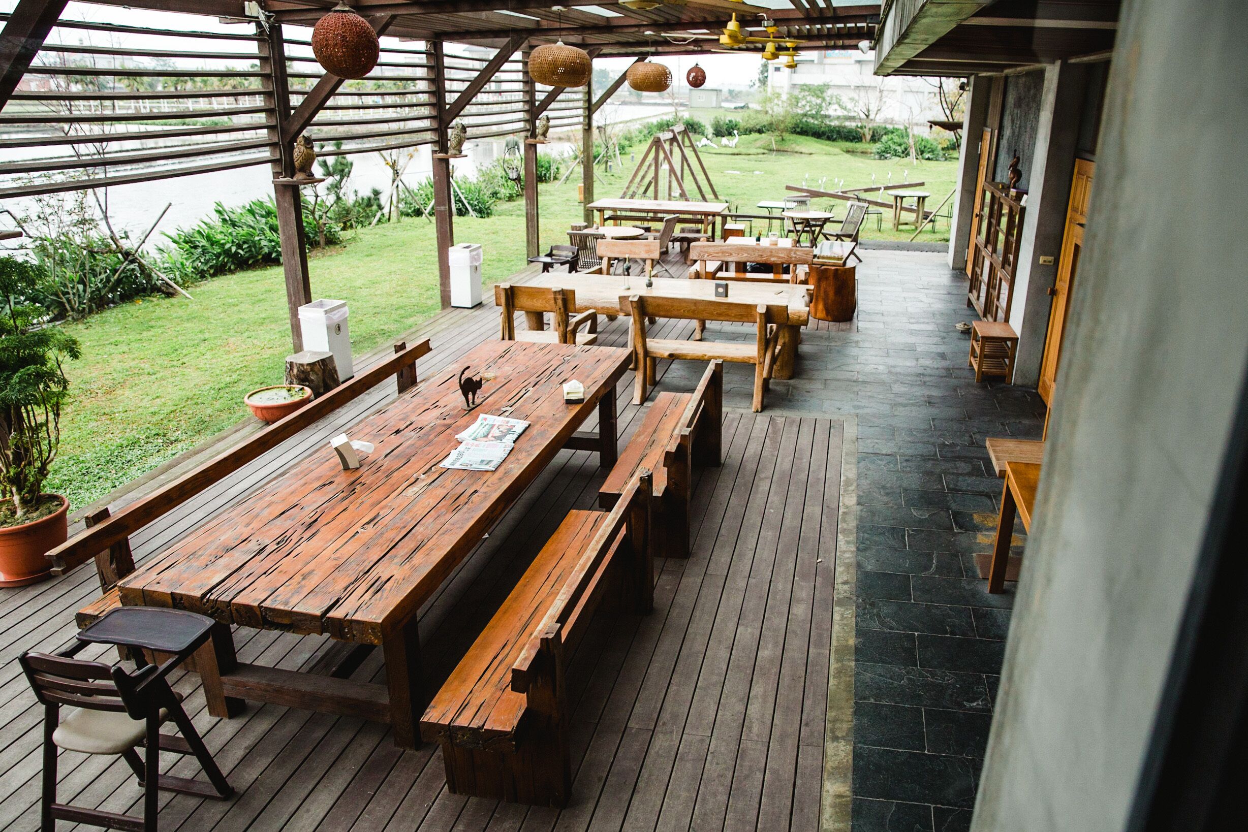 An outdoor wooden deck with picnic tables and benches, surrounded by greenery and a view of a body of water.