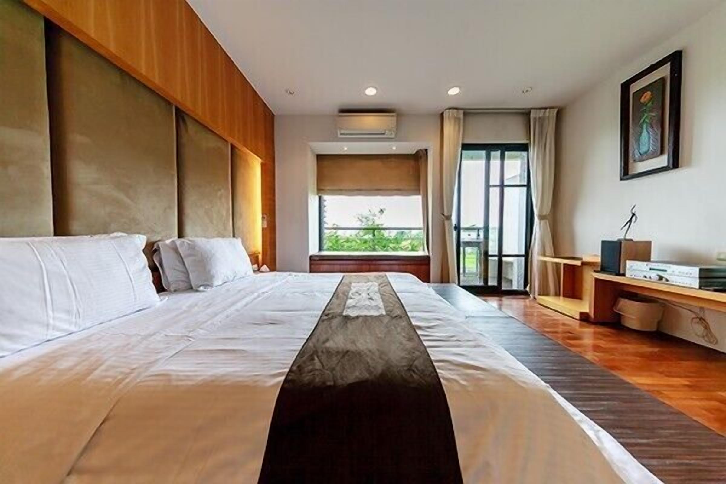 A hotel room with a large bed, a window with a view of greenery, a wooden headboard, and a desk with a lamp.