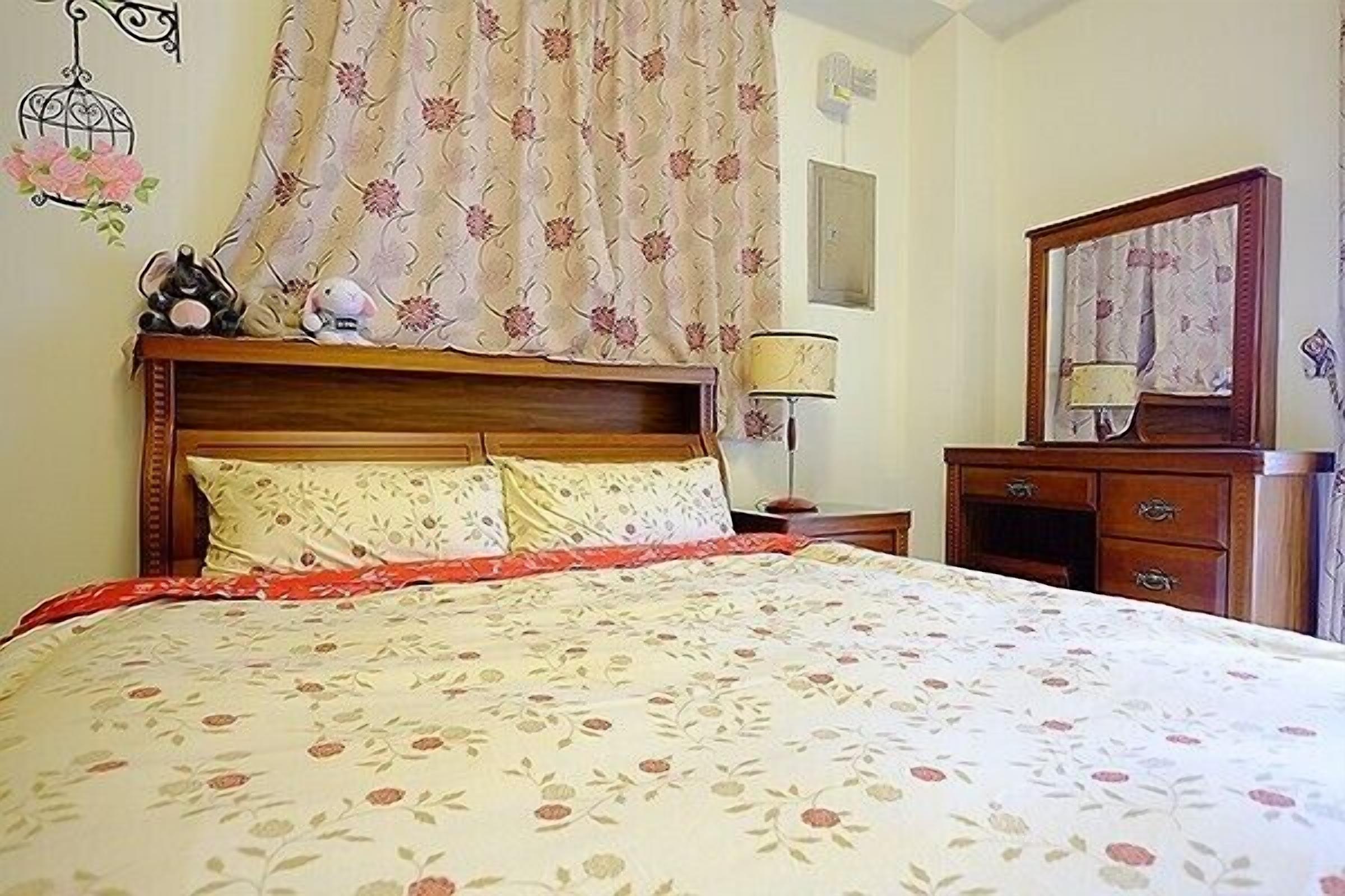 Una cama bien tendida con ropa de cama floral, un cabecero de madera, una mesita de noche con una lámpara y un aparador con un espejo.