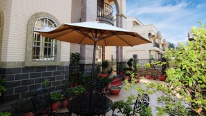 Une cour avec une table et des chaises sous un parasol, entourée de plantes en pot et d’un bâtiment doté d’un balcon.