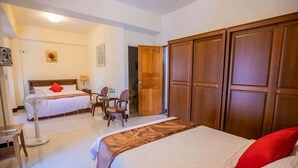 Une chambre d’hôtel avec un grand lit, un lit plus petit, un bureau avec des chaises, une armoire et un ventilateur.