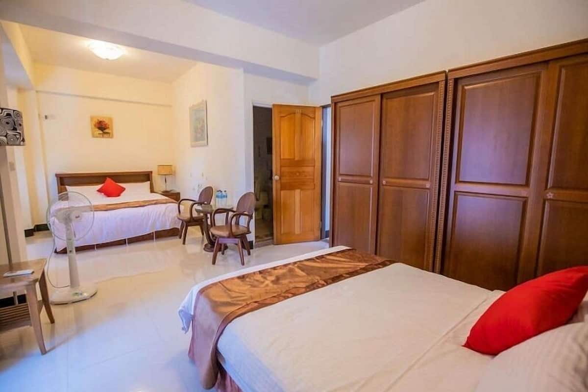 A hotel room with a large bed, a smaller bed, a desk with chairs, a wardrobe, and a fan.