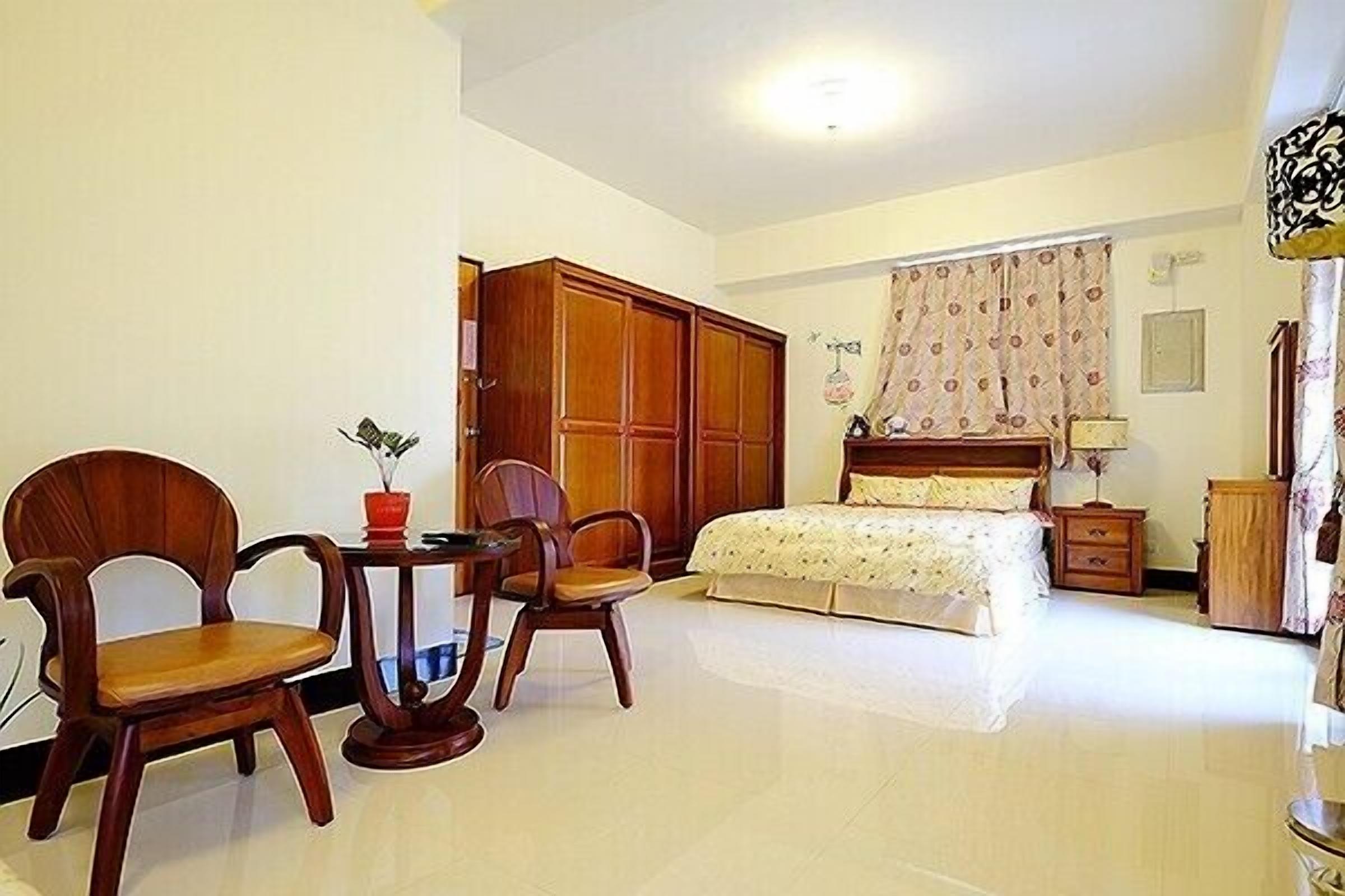 A bedroom with a bed, a wooden wardrobe, two wooden chairs, a small table with a red vase, and a nightstand.