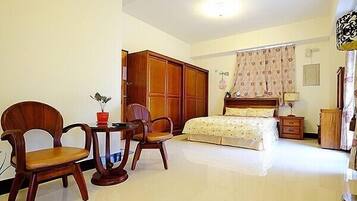 A bedroom with a bed, a wooden wardrobe, two wooden chairs, a small table with a red vase, and a nightstand.