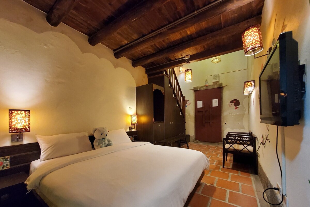 A hotel room with a large bed, wooden ceiling beams, a small table with a chair, and decorative wall sconces.