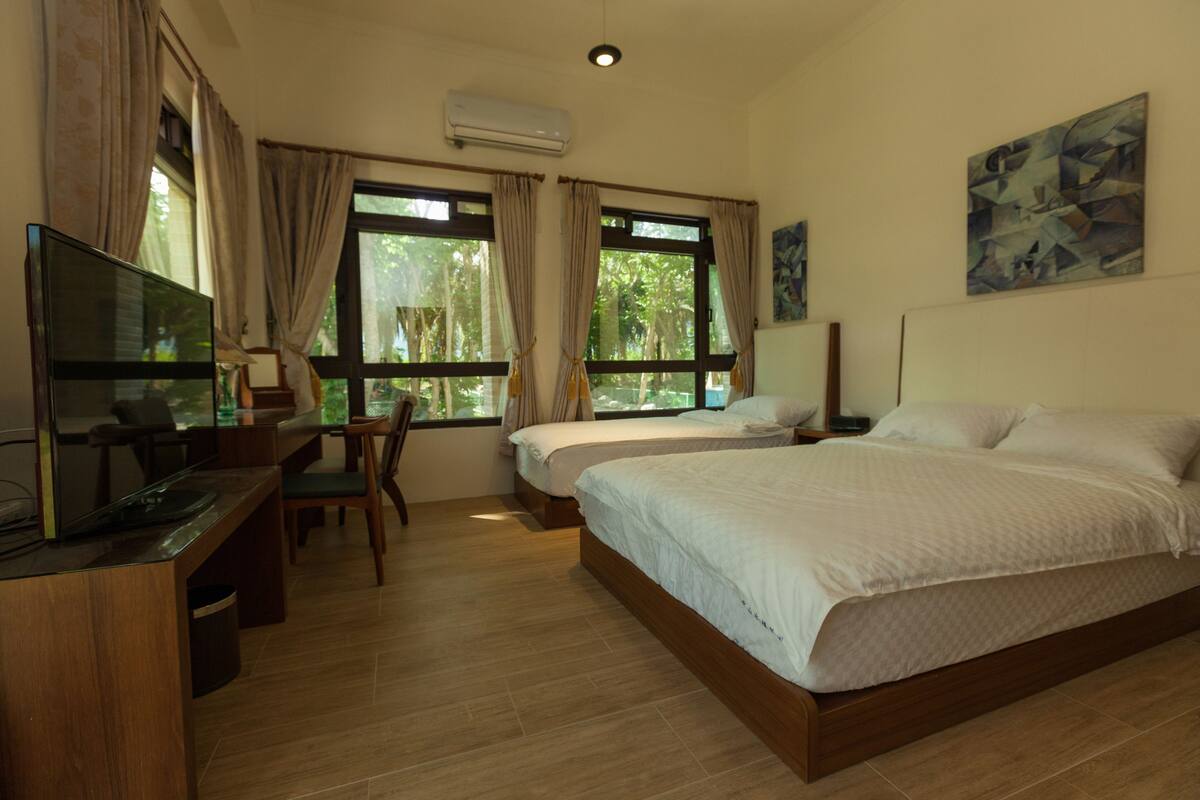 A hotel room with a bed, a desk, a chair, and a television. There are windows with curtains and a painting on the wall.