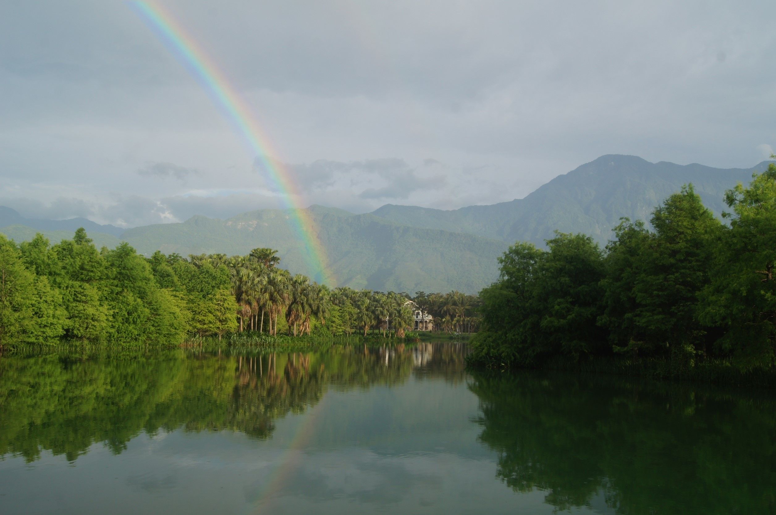 A tranquil lake with a rainbow, surrounded by lush greenery and mountains.