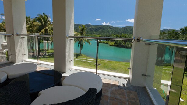 A balcony with a glass railing, white cushioned seating, and a view of a turquoise lake and greenery. - Villa of Permaculture (Shoufeng)