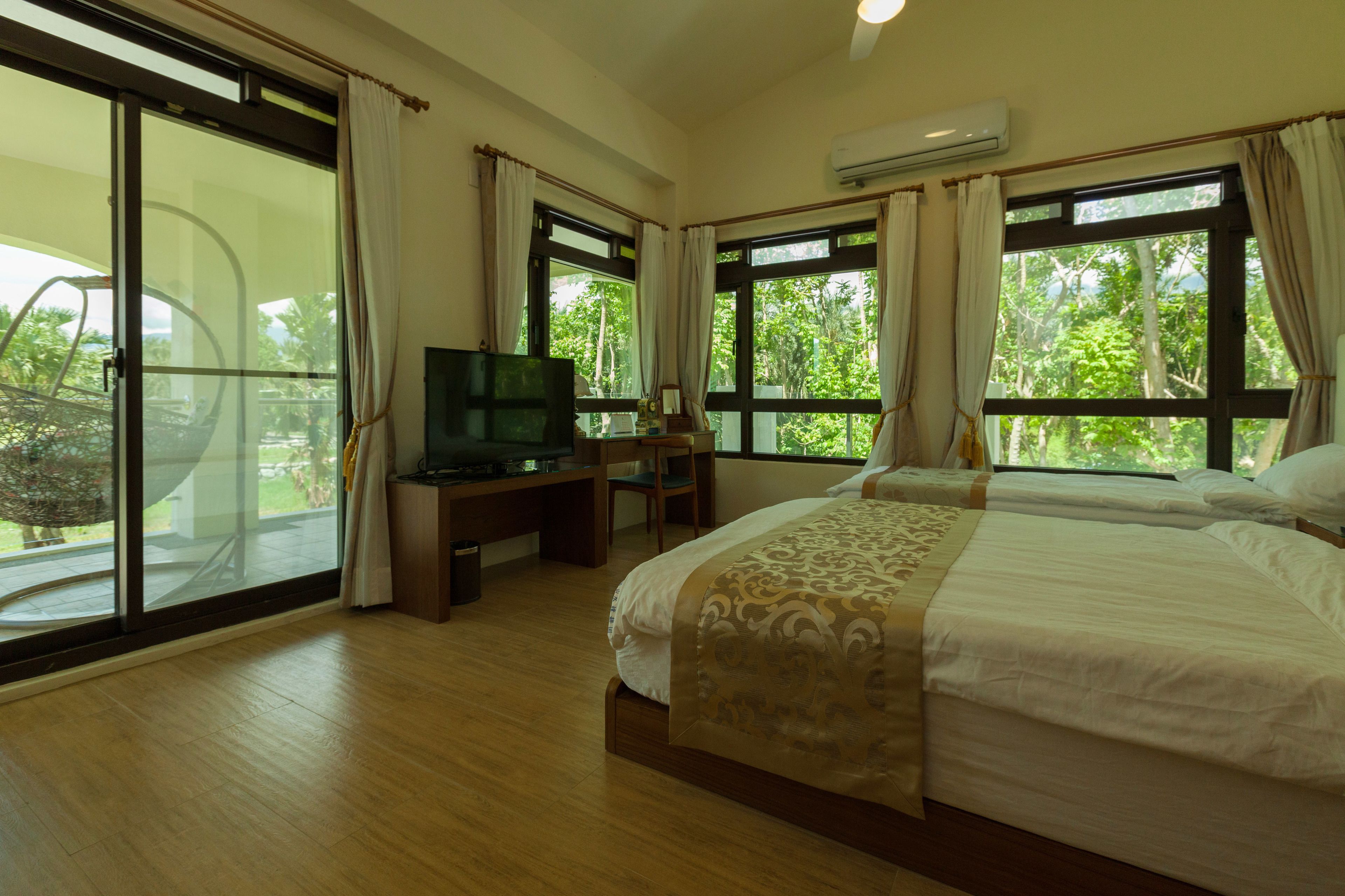 Habitación de hotel con cama, escritorio con televisor y grandes ventanales que dan a zonas verdes.