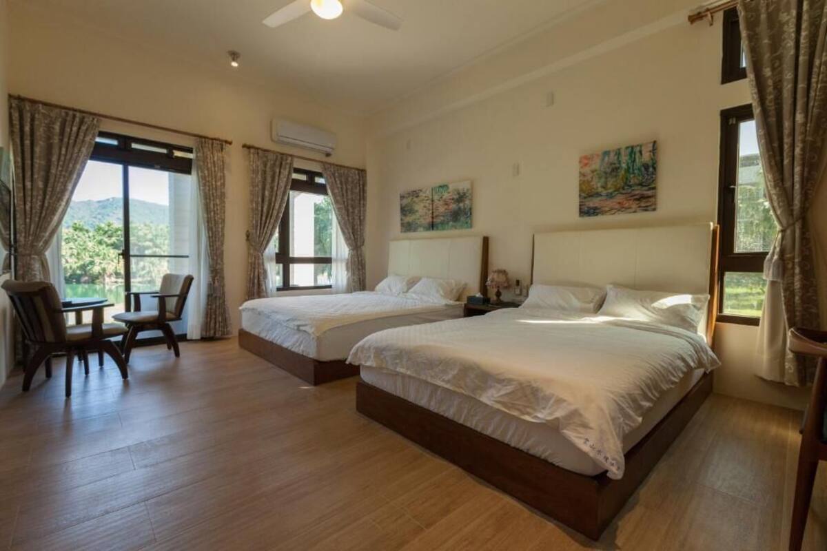 A hotel room with two single beds, a wooden floor, a dining area with a table and chairs, and large windows with curtains.