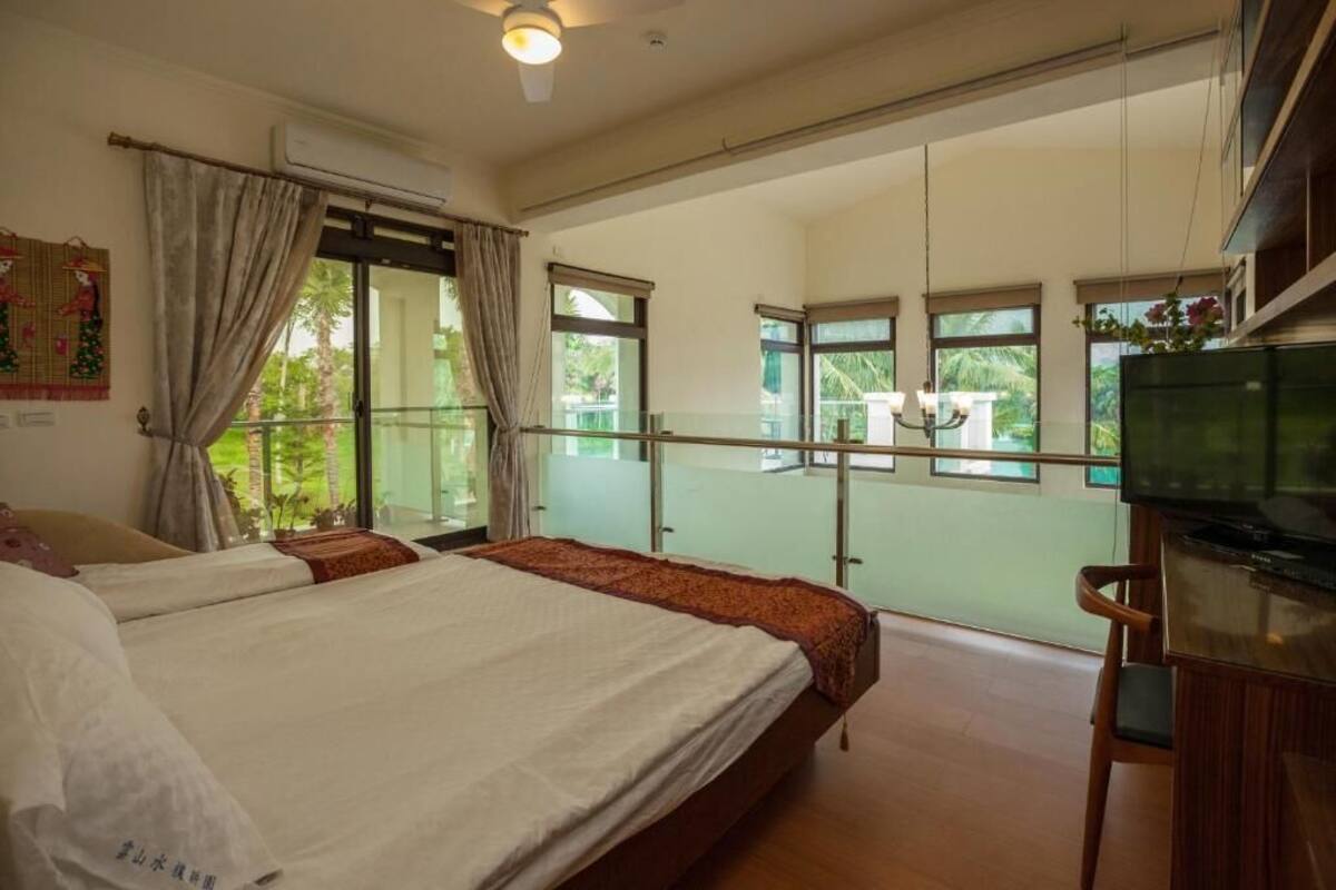 A bedroom with two beds, a balcony with a glass railing, a television, and a ceiling fan.
