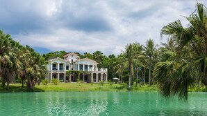 A large, two-story house with a symmetrical facade, surrounded by a body of water and palm trees. - Villa of Permaculture (Shoufeng)