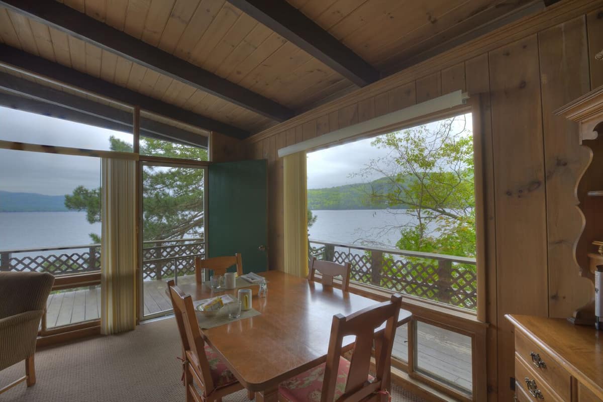 A wooden dining area with a table and chairs, large windows with a view of a lake and trees, and a wooden ceiling.