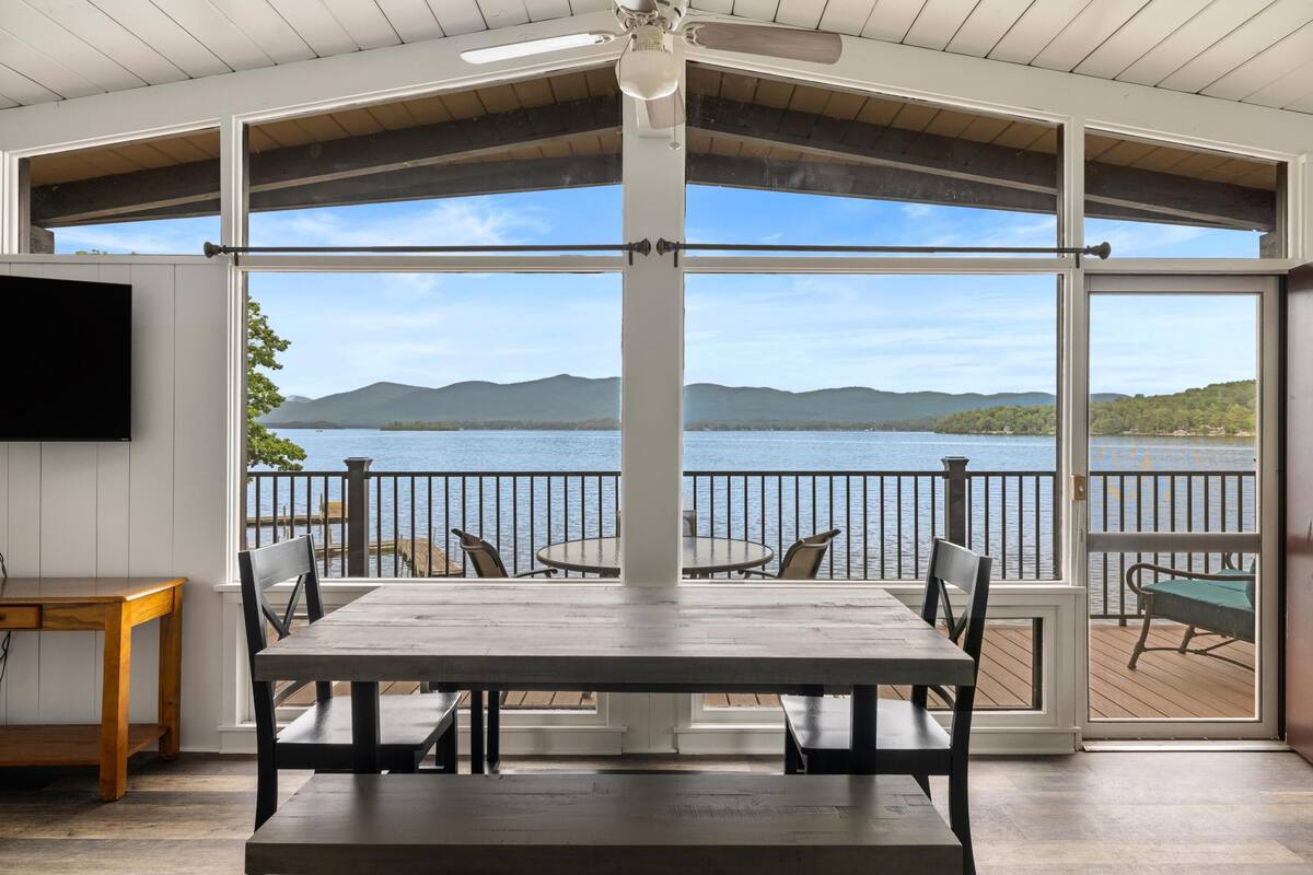 A dining area with a table and chairs, a television mounted on the wall, and a view of a lake and mountains through large windows.