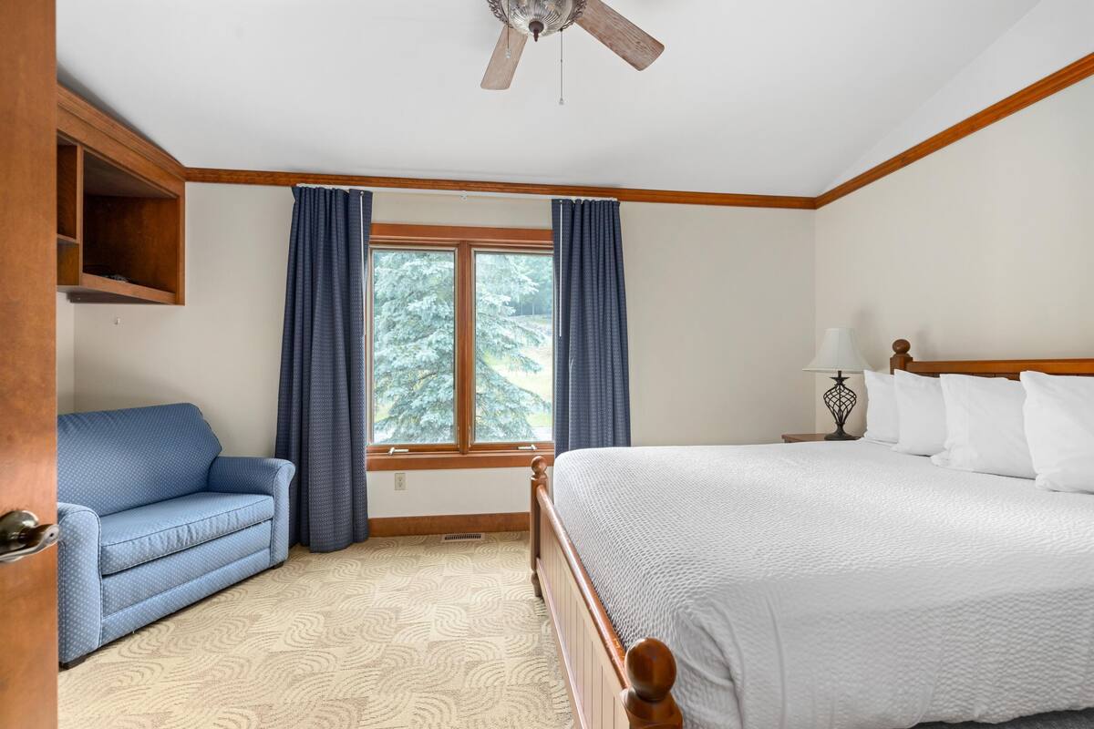 A bedroom with a bed, a blue sofa, a window with curtains, a ceiling fan, and a wooden shelf.