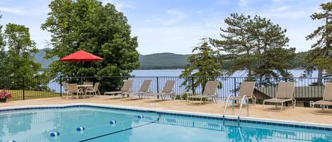A swimming pool with lounge chairs and a red umbrella, surrounded by trees and a view of a lake.