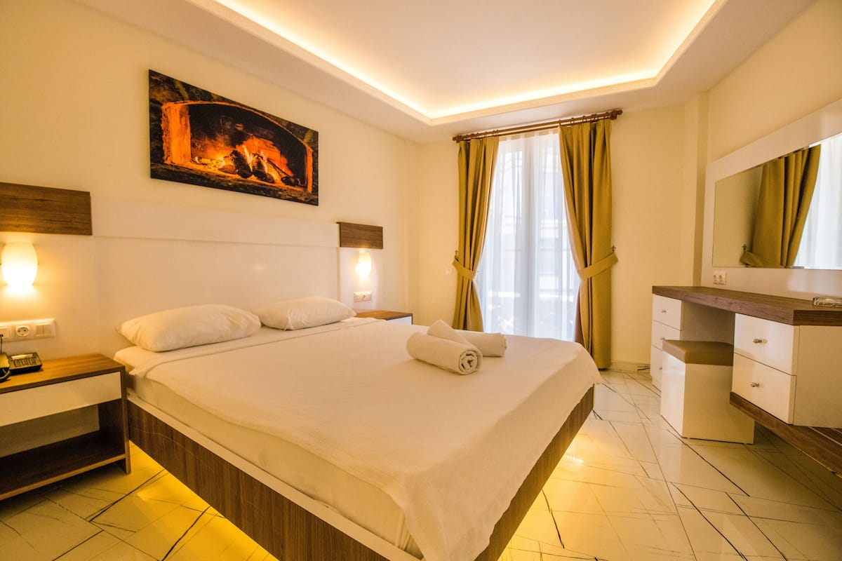 Une chambre d’hôtel moderne avec un grand lit, des lampes de chevet, un tableau, une coiffeuse avec un miroir et une fenêtre avec des rideaux.