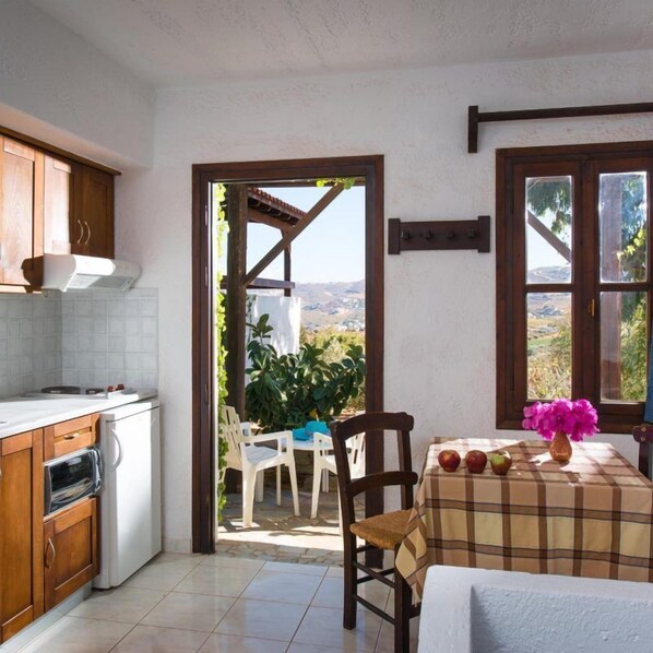 A kitchen with a dining area, a view through an open door, and a table with apples and flowers. - Ambelos (Malevizi)
