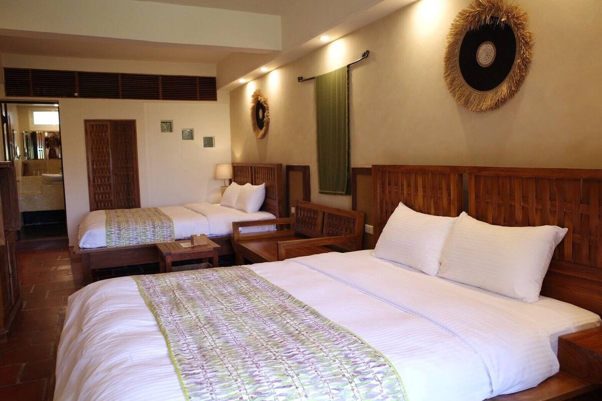 A hotel room with two beds, wooden headboards, a large bedspread, a bedside table with a lamp, and a wall-mounted mirror.