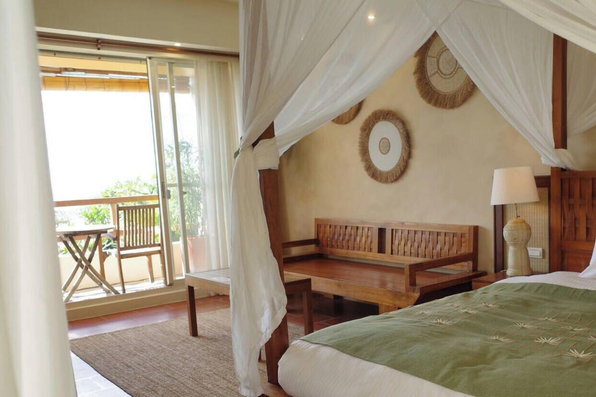 A bedroom with a bed, a wooden bench, a nightstand, a lamp, and a balcony with a table and chairs.