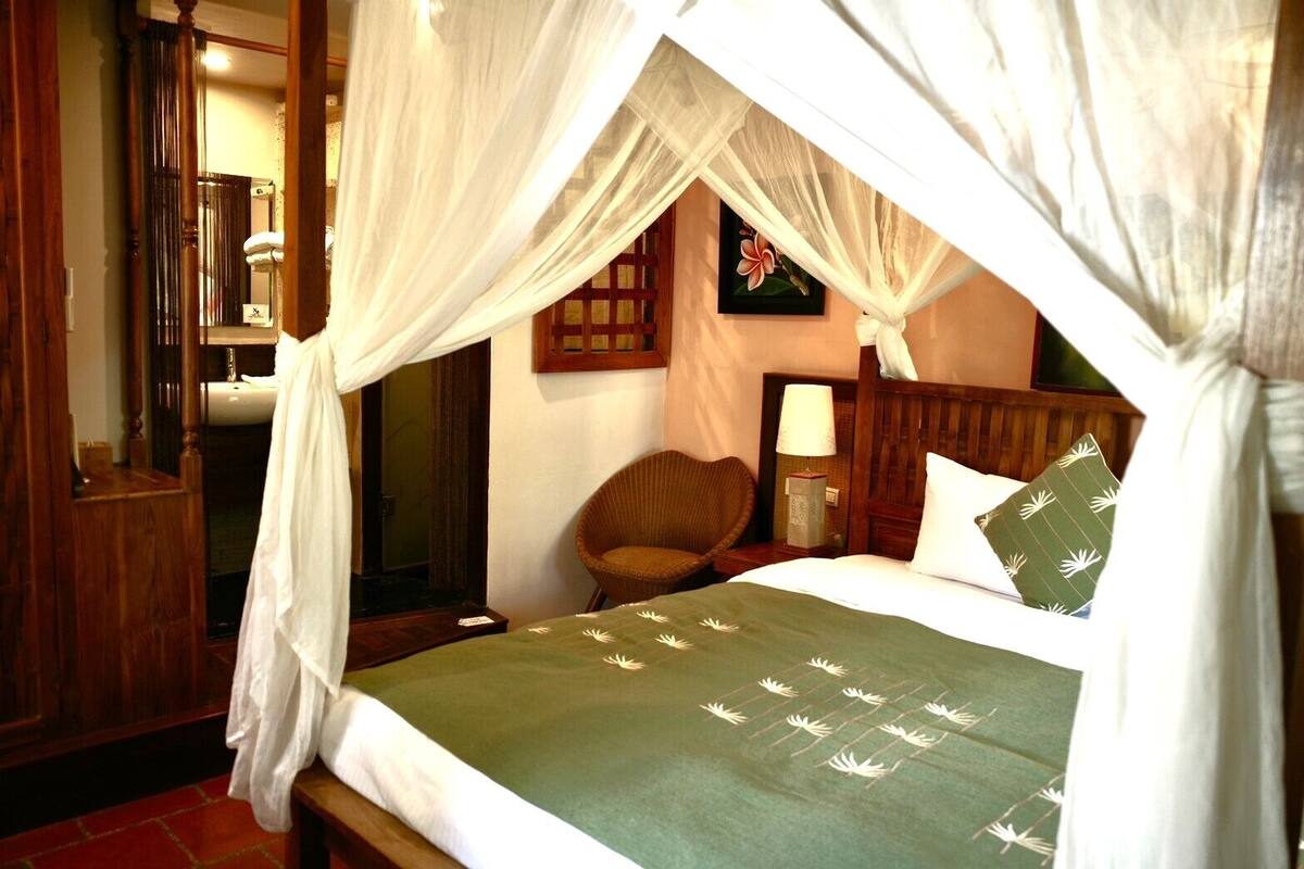 A bedroom with a canopy bed, a wooden headboard, a bedside table with a lamp, and a chair.
