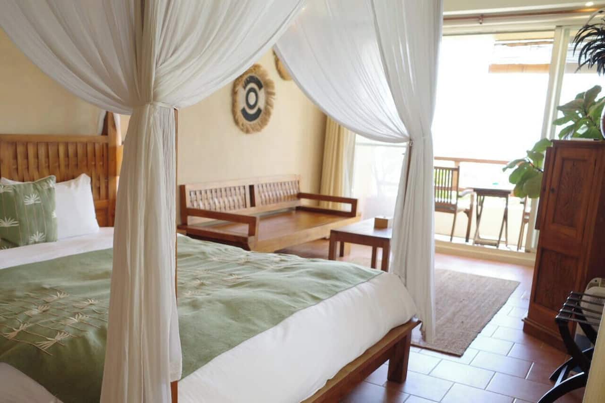 A bed with a canopy, a wooden headboard, and a green bedspread. There is a wooden bench and a plant in the corner.