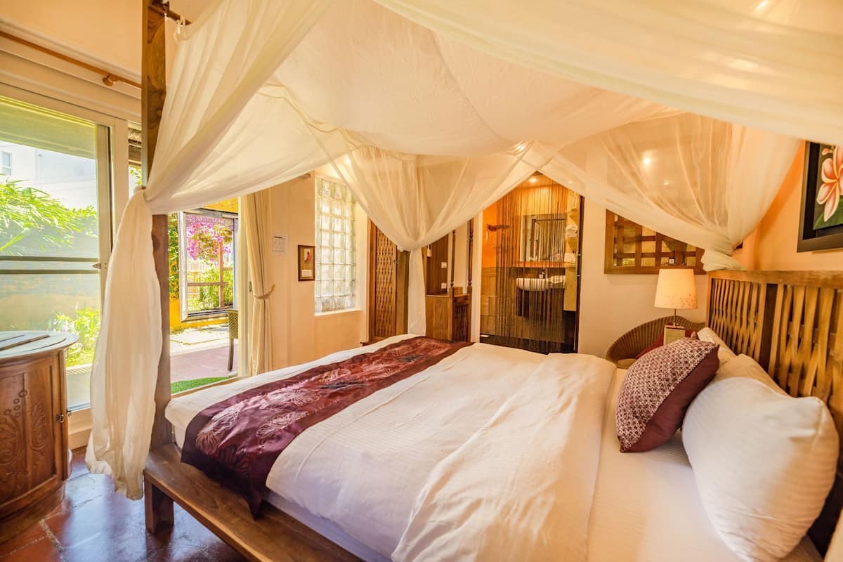 A bed with a canopy, a bedside table, a window with curtains, and a view of greenery outside.