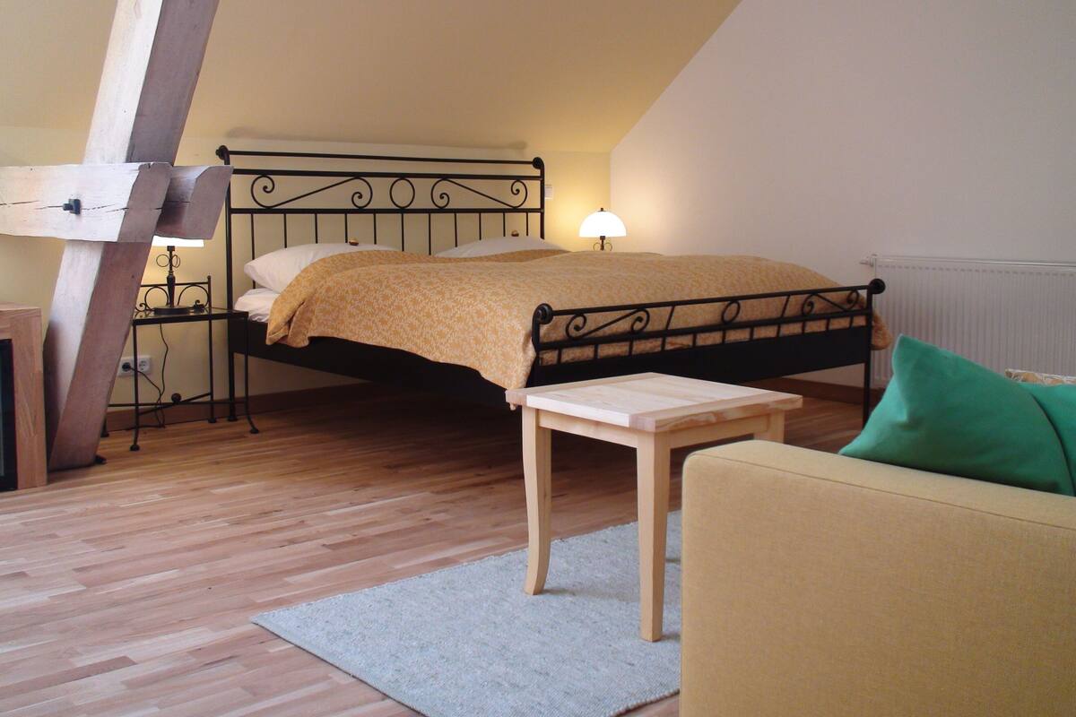 A bedroom with a wooden floor, a bed with a headboard, a bedside table, a lamp, and a green pillow on a sofa.