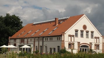 A large, two-story building with a red-tiled roof, multiple windows, and a central circular window. There are outdoor tables and umbrellas, suggesting a patio area.
