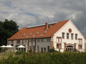 A large, two-story building with a red-tiled roof, multiple windows, and a central circular window. There are outdoor tables and umbrellas, suggesting a patio area.