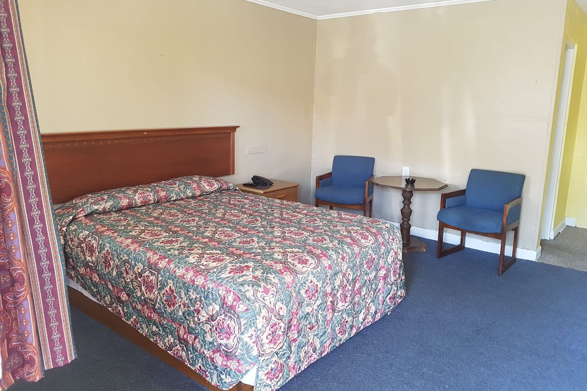 Hotellihuone, jossa on sänky, kaksi tuolia, pieni pöytä ja yöpöytä.