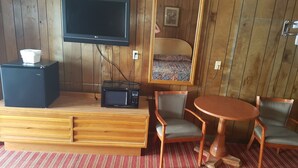 Una habitación compacta con piso de madera, un televisor de pantalla plana montado en la pared, un microondas negro pequeño, un mueble de madera con una caja de pañuelos, una mesa de madera redonda y dos sillas.