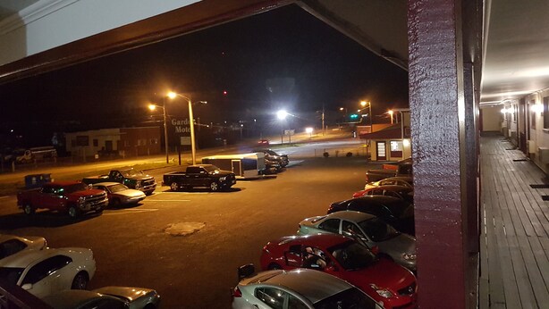 Un estacionamiento nocturno con varios autos estacionados, un edificio con un letrero que dice 'Garden Motel' y una calle con semáforos y una intersección en el fondo.