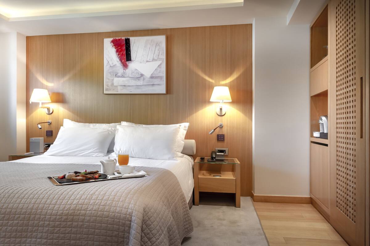 Habitación de hotel con una cama grande, dos lámparas de noche, una mesita de noche con teléfono y lámpara, cabecero de madera, un cuadro y un armario de madera.