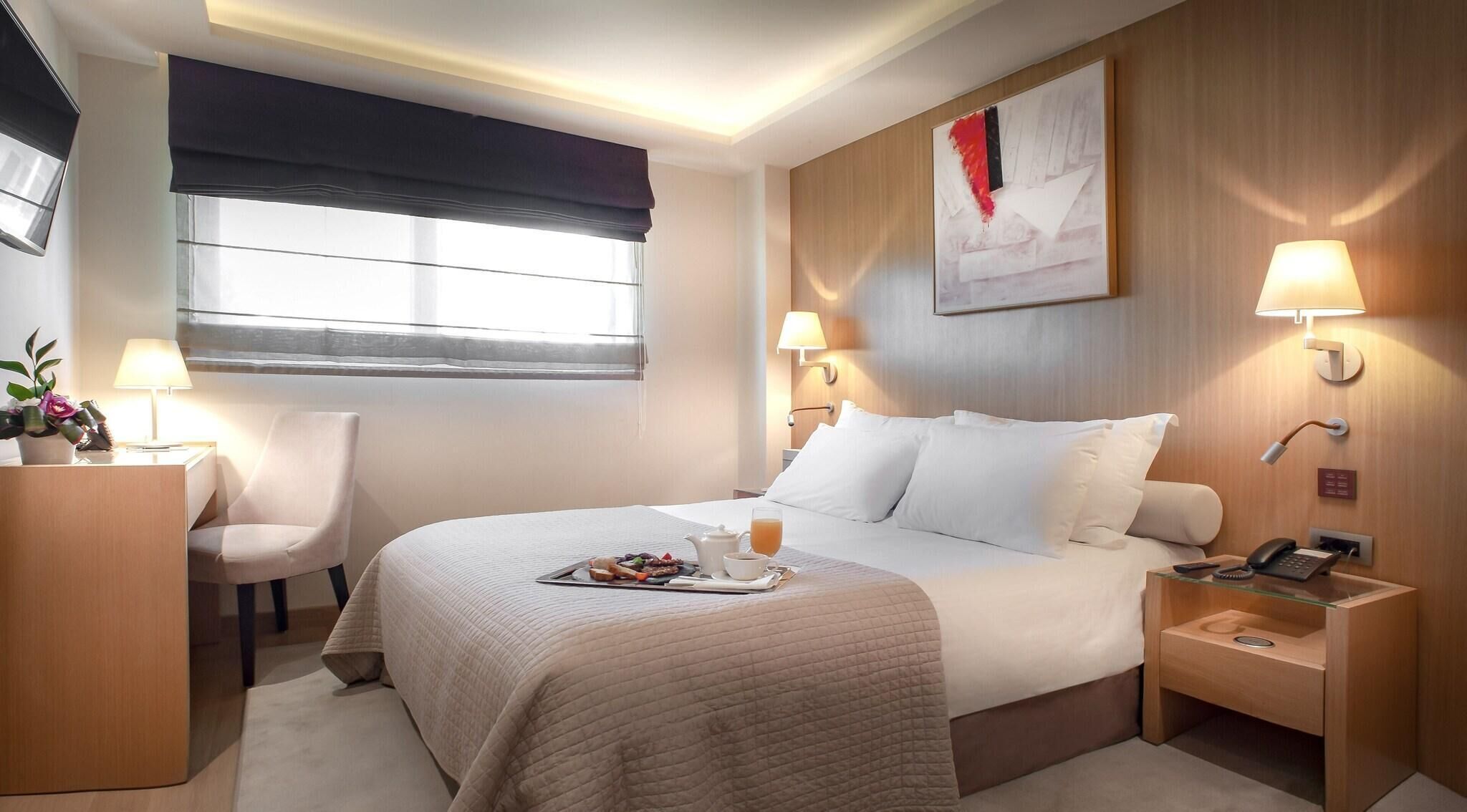 A hotel room with a bed, bedside tables, a desk, a chair, a window with a view, and a painting on the wall.