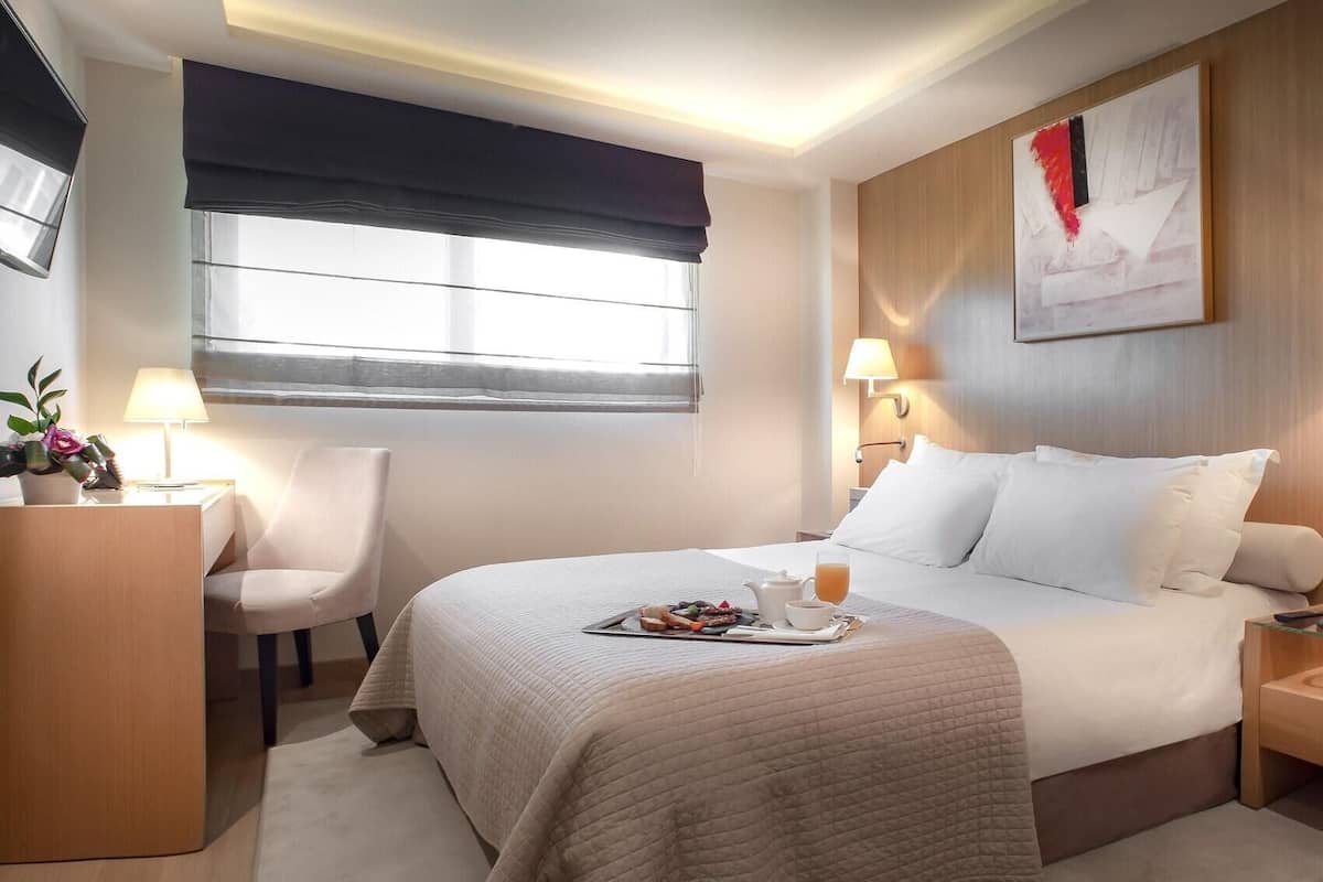 A hotel room with a bed, bedside tables, a desk, a chair, a window with a view, and a painting on the wall.