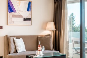 A hotel room with a beige sofa, a glass coffee table with a wine bottle and glass, a lamp, and a view of trees through a window.