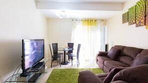 Een woonkamer met een flatscreen televisie op een glazen voetstuk, een bruine bank, een groen tapijt, en een eethoek met een tafel en stoelen.