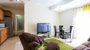 Een woonkamer met een bank, een televisie op een glazen bijzettafeltje, en een eethoek met stoelen en een tafel.