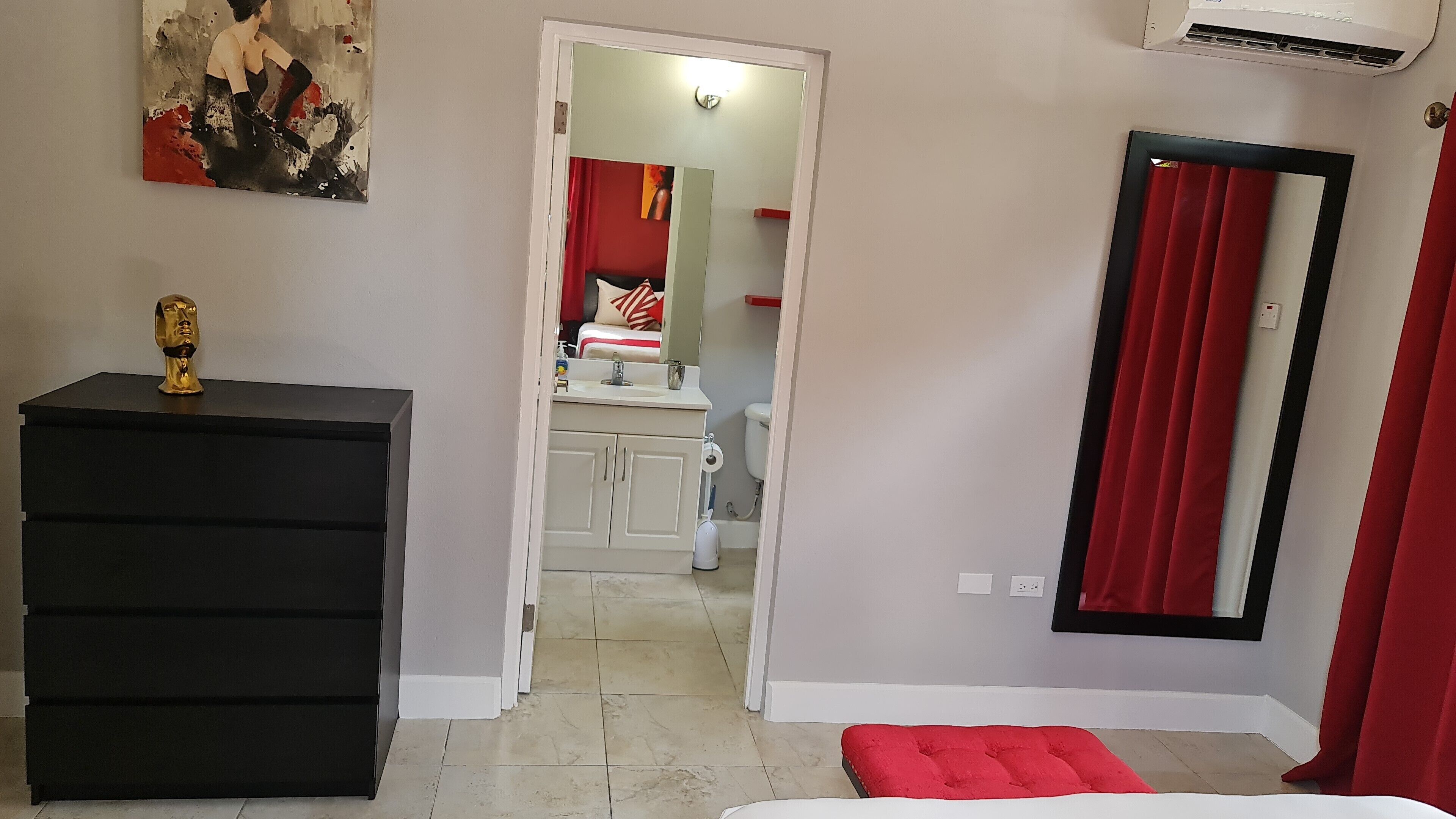 Ein Schlafzimmer mit einem schwarzen Kommode, einem roten Hocker, einem Bett und einem durchgehenden Türspalt sichtbaren Badezimmer.