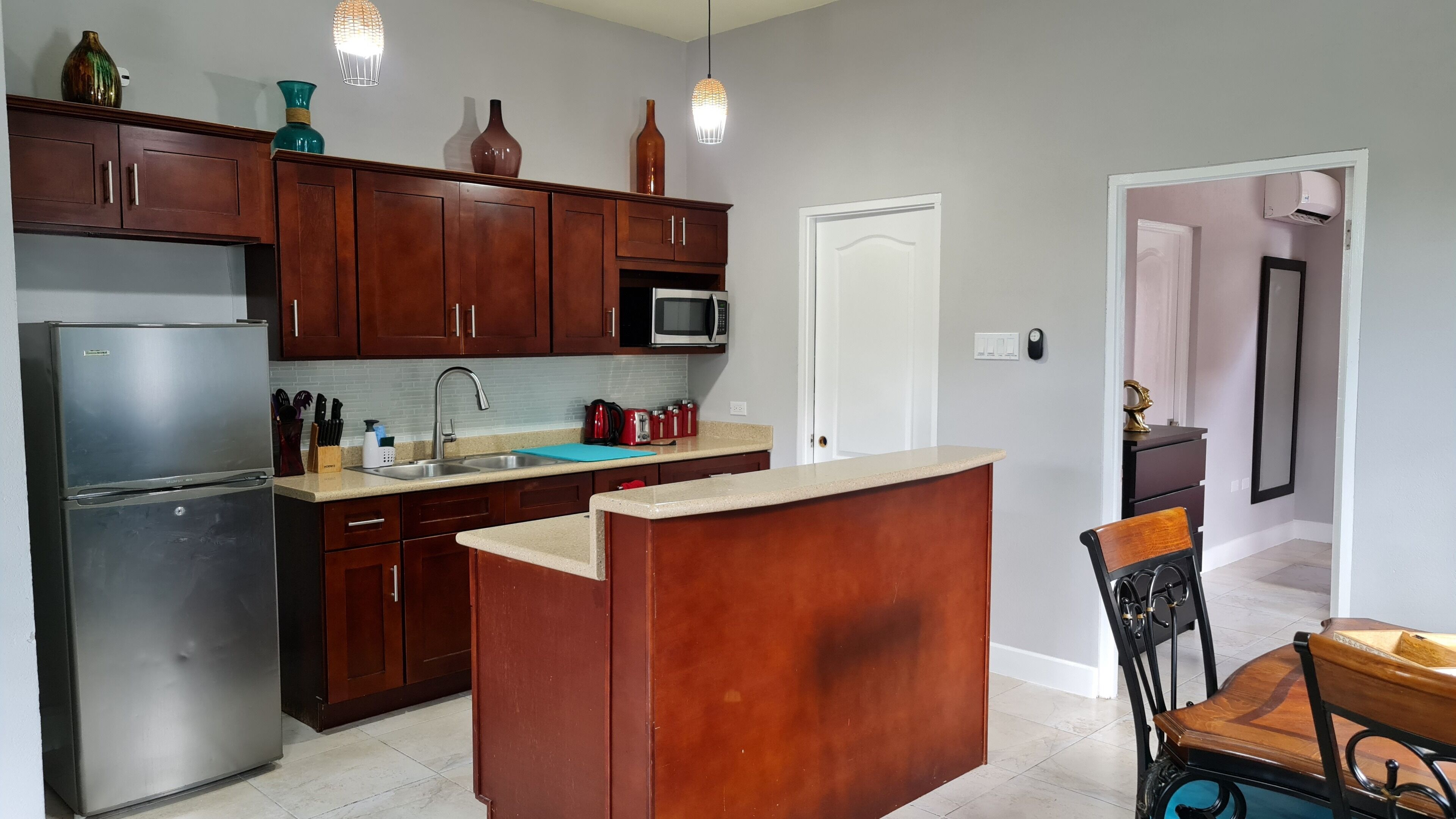 A modern kitchen with dark wooden cabinets, a stainless steel refrigerator, a microwave, and a sink. There are hanging pendant lights, a small island with a sink, and a chair with a wooden backrest.