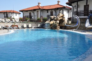 A swimming pool with lounge chairs, a rock waterfall feature, and a building with red-tiled roofs in the background.