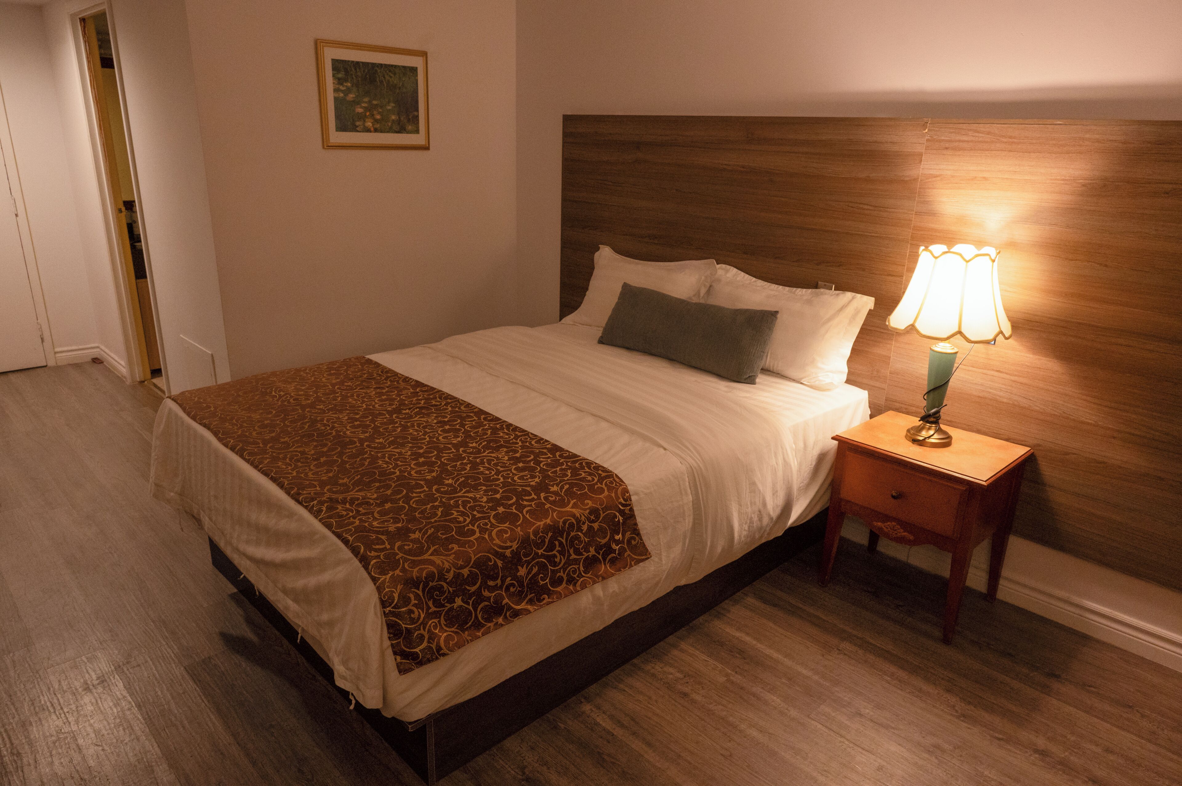 A hotel room with a bed, a bedside lamp, a wooden headboard, a patterned throw, and a framed picture on the wall.