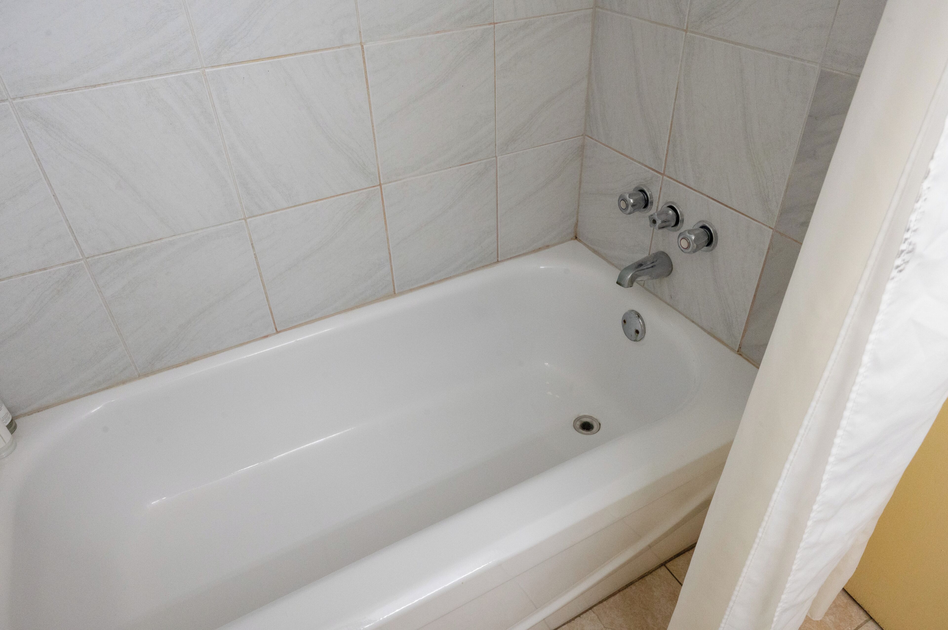 Una bañera blanca con dos llaves y un desagüe, frente a una pared azulejada.